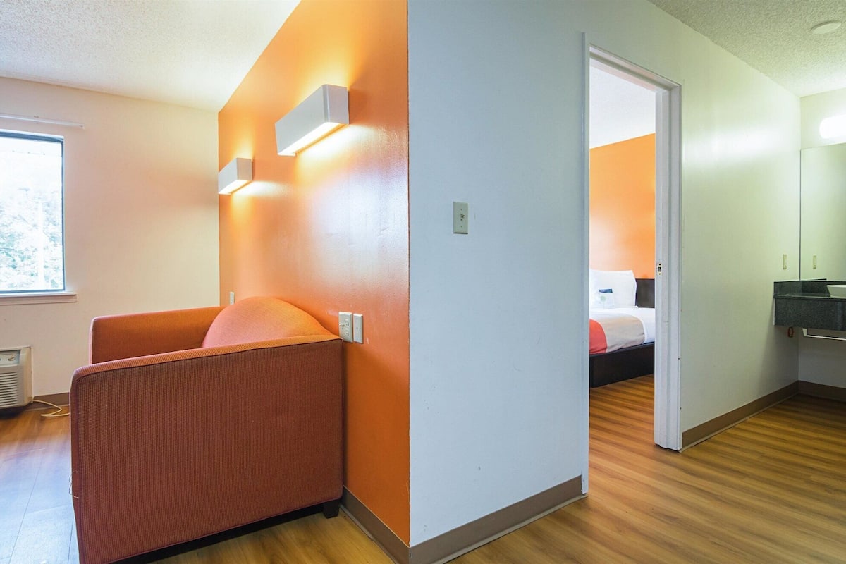 A hotel room with a wooden floor, a red upholstered chair, a bed with white bedding, and a bathroom with a mirror and sink.