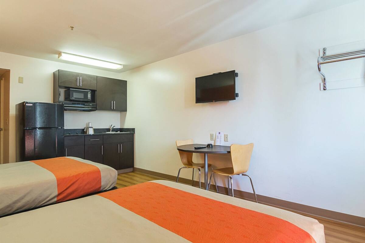 A hotel room with two beds, a kitchenette, a TV, and a small table with chairs.