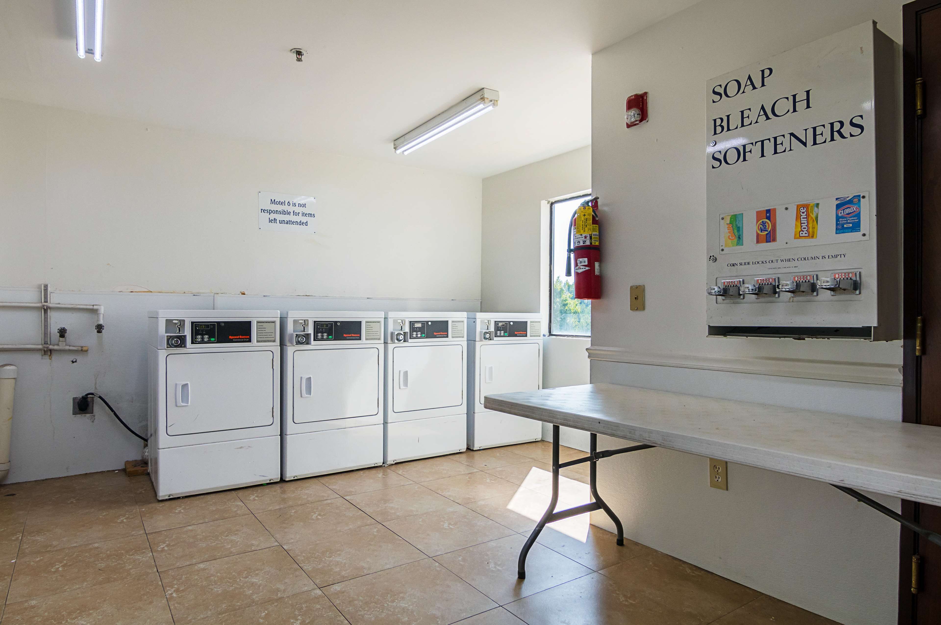 A laundry room with four white washers and dryers, a table, and a wall sign advertising soap, bleach, and softeners.