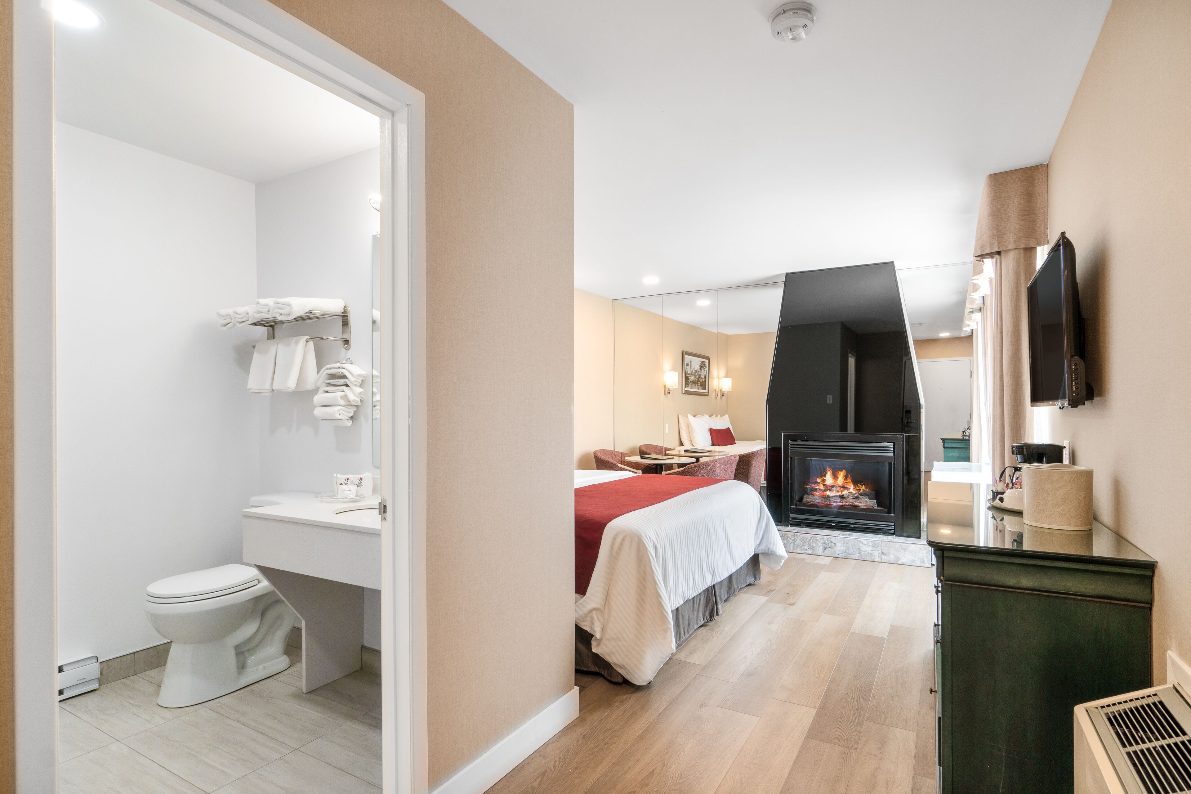 Una habitación de hotel moderna con baño, una cama con una colcha roja, una chimenea y un televisor montado en la pared.