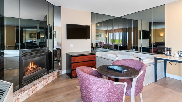 A modern living room with a fireplace, a dining area with chairs, a kitchen with a large mirror, and a view of the outdoors. - Hotel Bromont (Bromont)