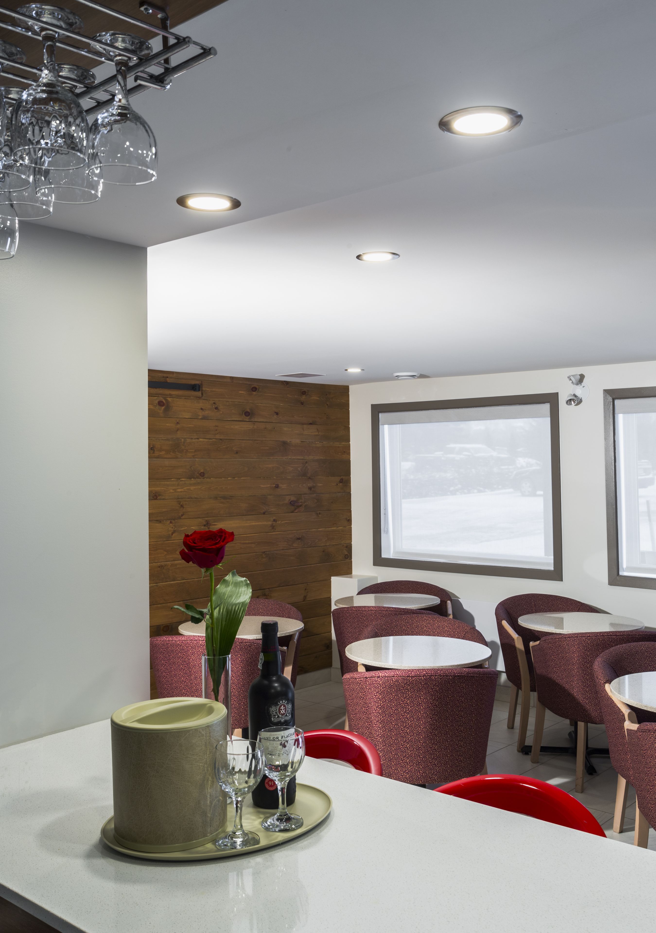 A restaurant interior with round tables and chairs, a wooden accent wall, and a large window.
