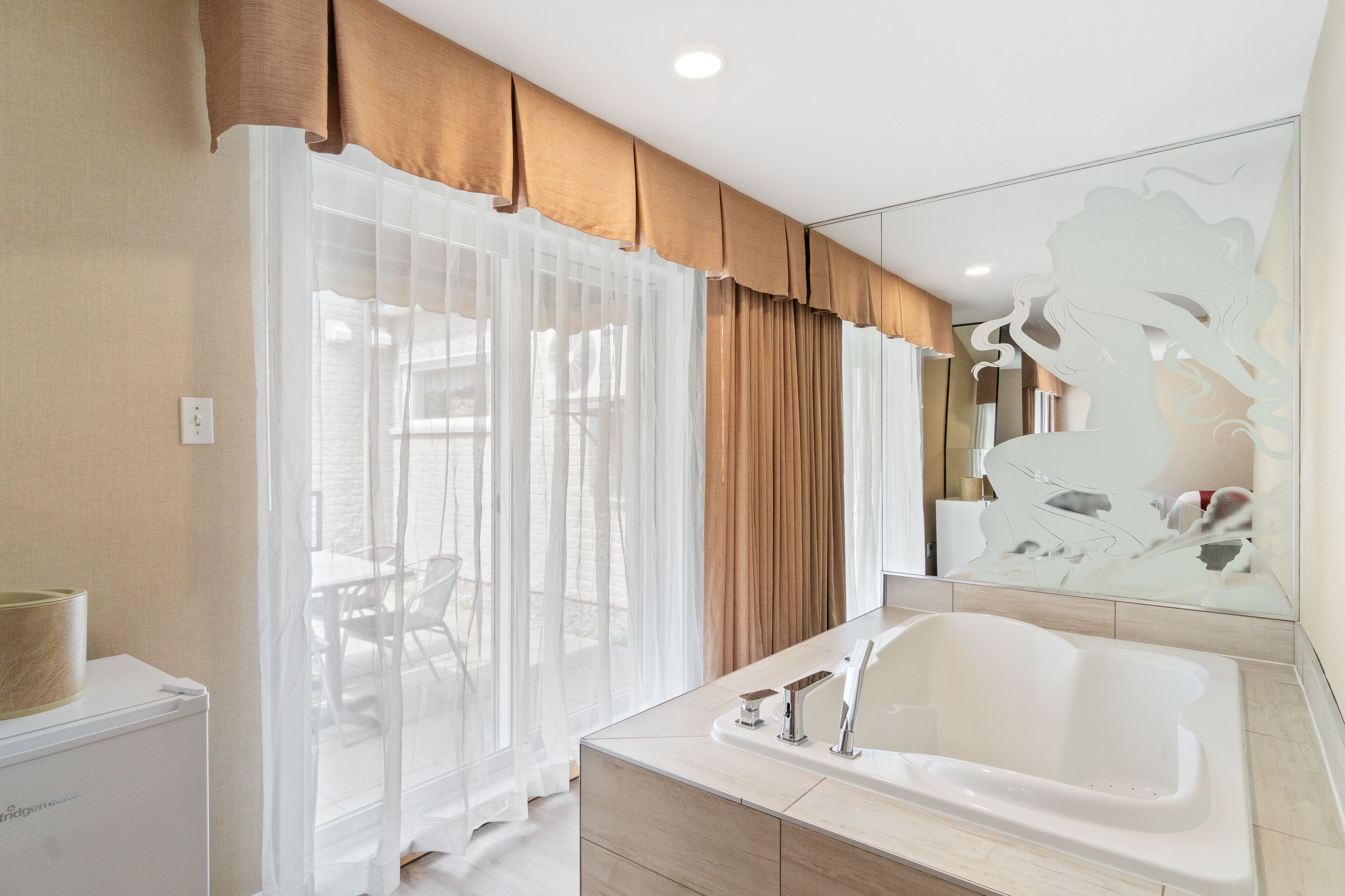 Een moderne badkamer met een groot bad, een spiegel met decoratieve lijst, en uitzicht op het buitengebied door dunne gordijnen.