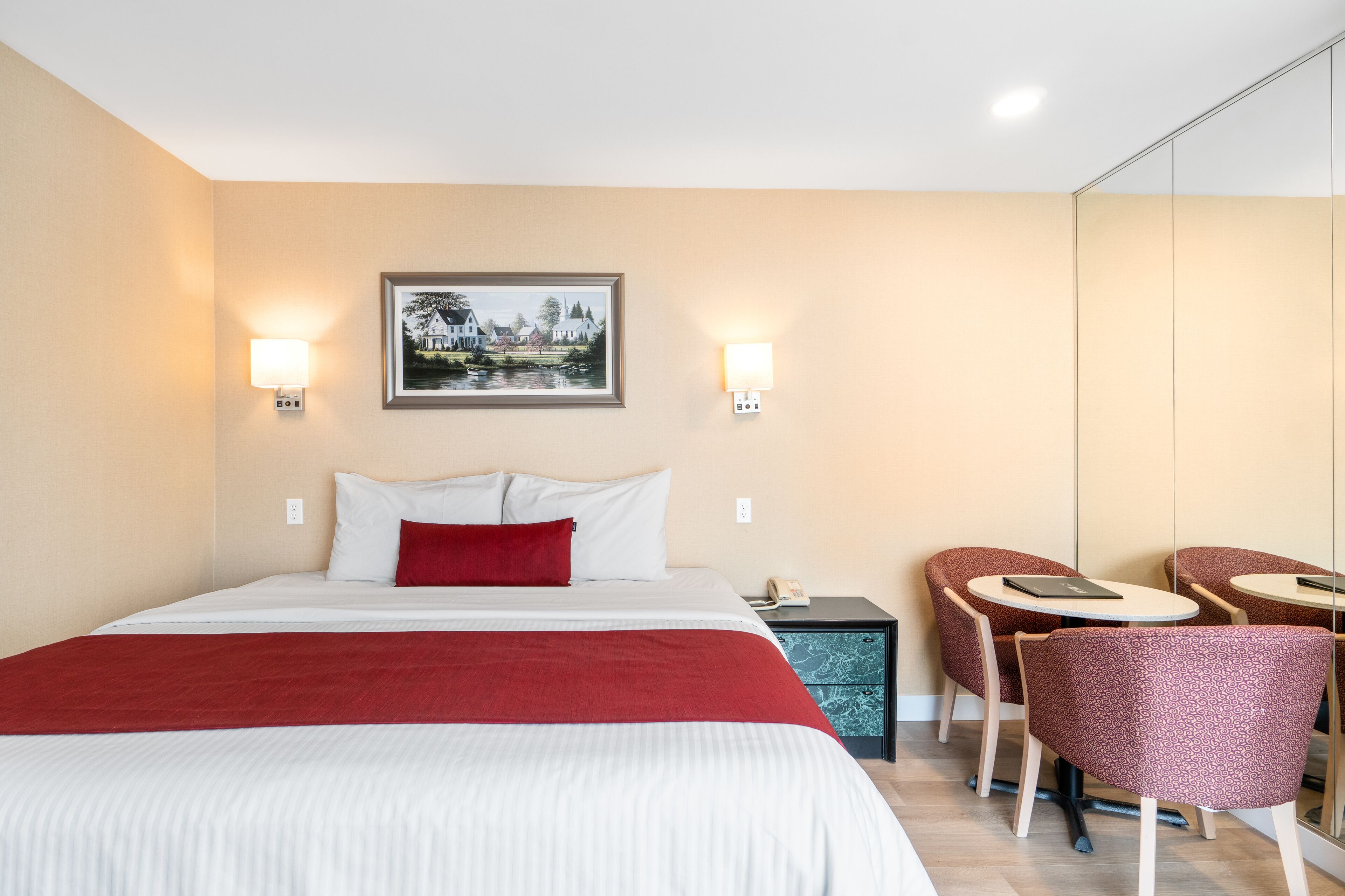 Habitación de hotel con cama, dos lámparas de noche, un cuadro en la pared, una mesita con una silla y un espejo.