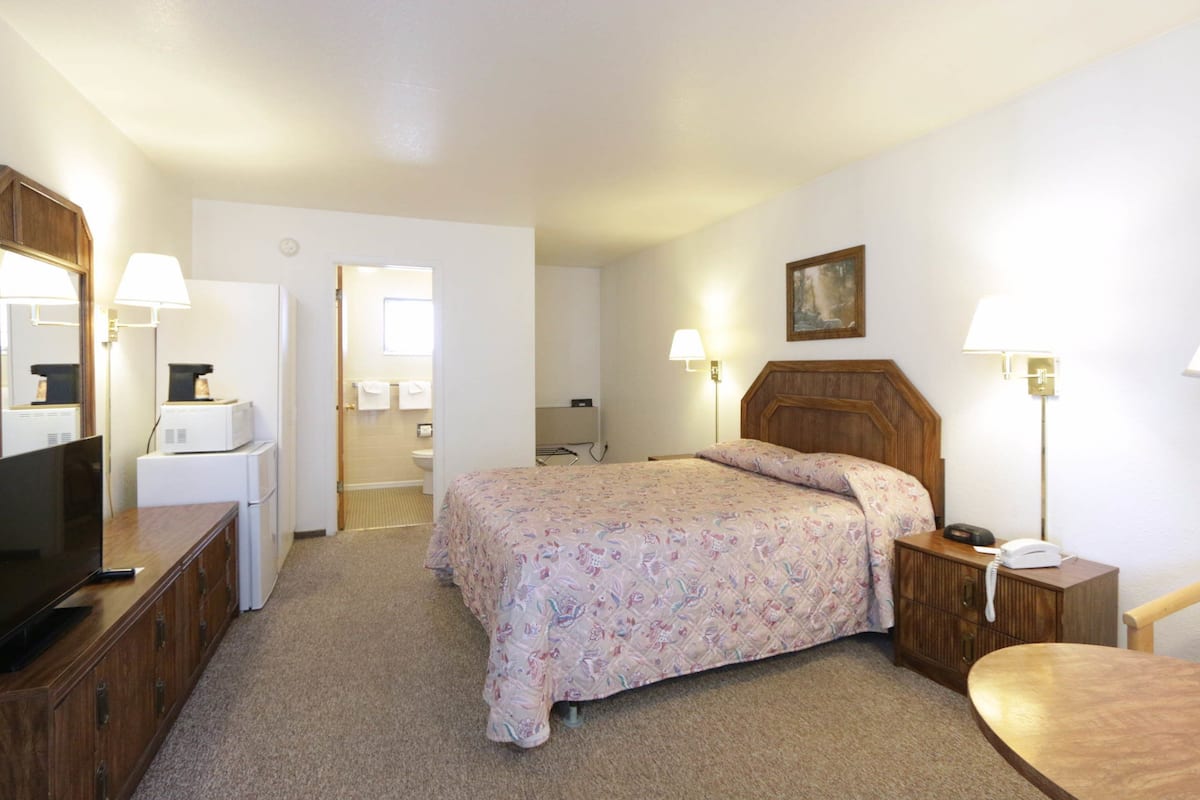 Habitación de hotel con cama, escritorio con televisor, mesita de noche con lámpara y baño visible a través de una puerta abierta.