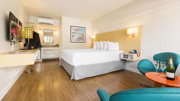 Habitación de hotel con cama, escritorio, silla, una mesita con una botella y vasos, y una lámpara de pared.
