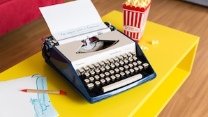 Una máquina de escribir sobre una mesa amarilla con un papel, una pluma roja y un recipiente para palomitas.
