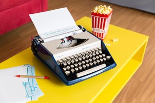 A typewriter on a yellow table with a piece of paper, a red pen, and a popcorn container.