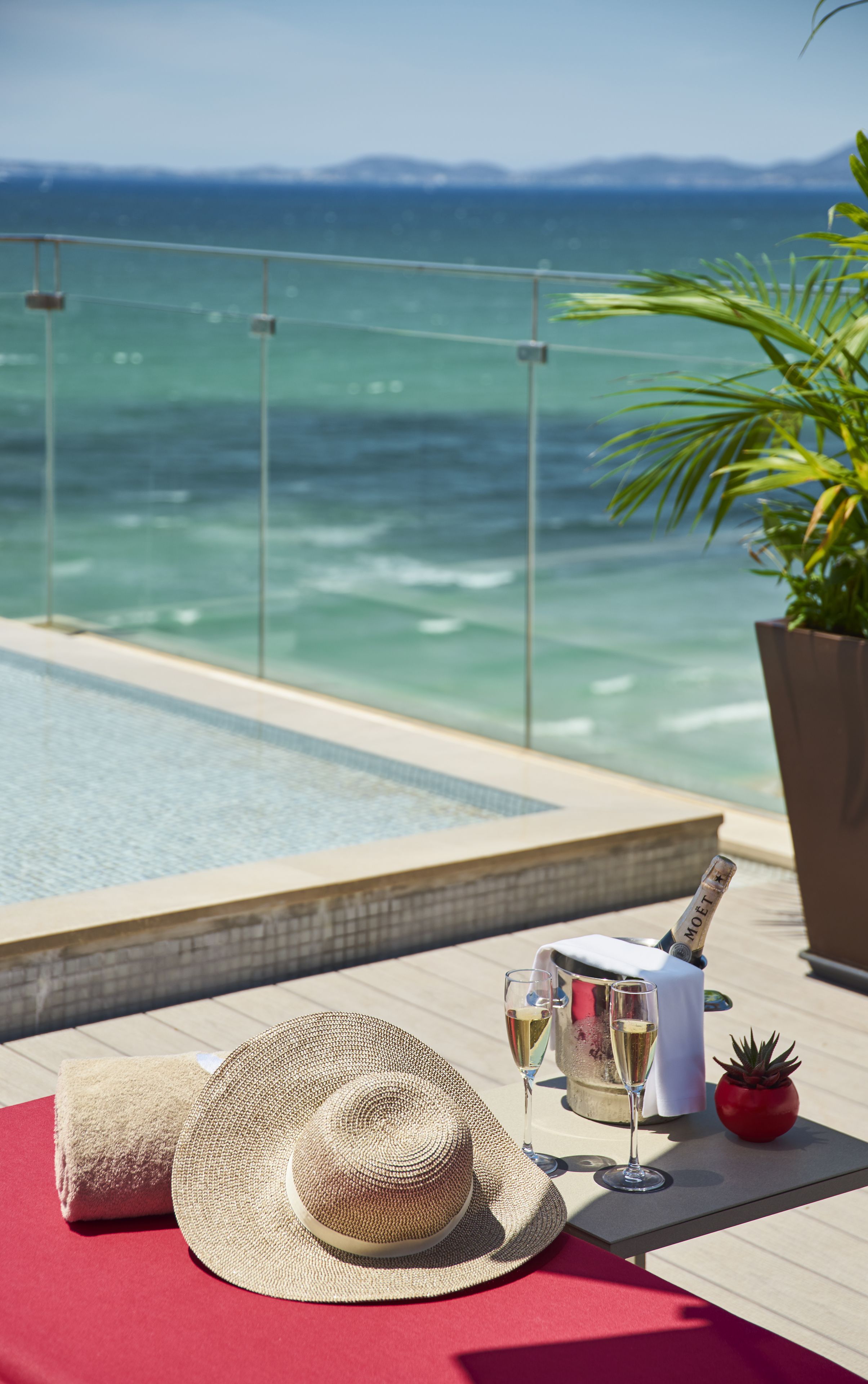 A poolside setting with a clear view of the ocean, a bottle of champagne, and a straw hat on a red mat.