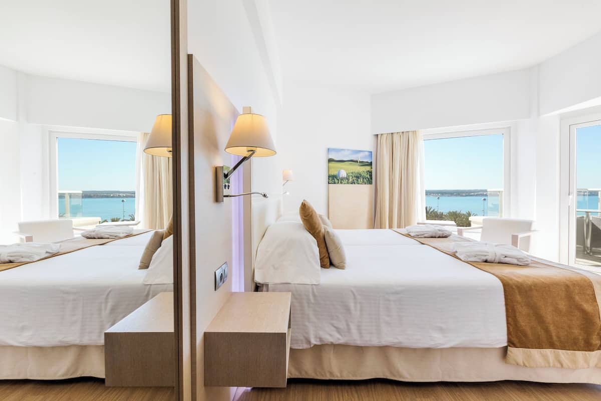 A hotel room with two beds, a large window with a view of the sea, a bedside table, and a lamp.