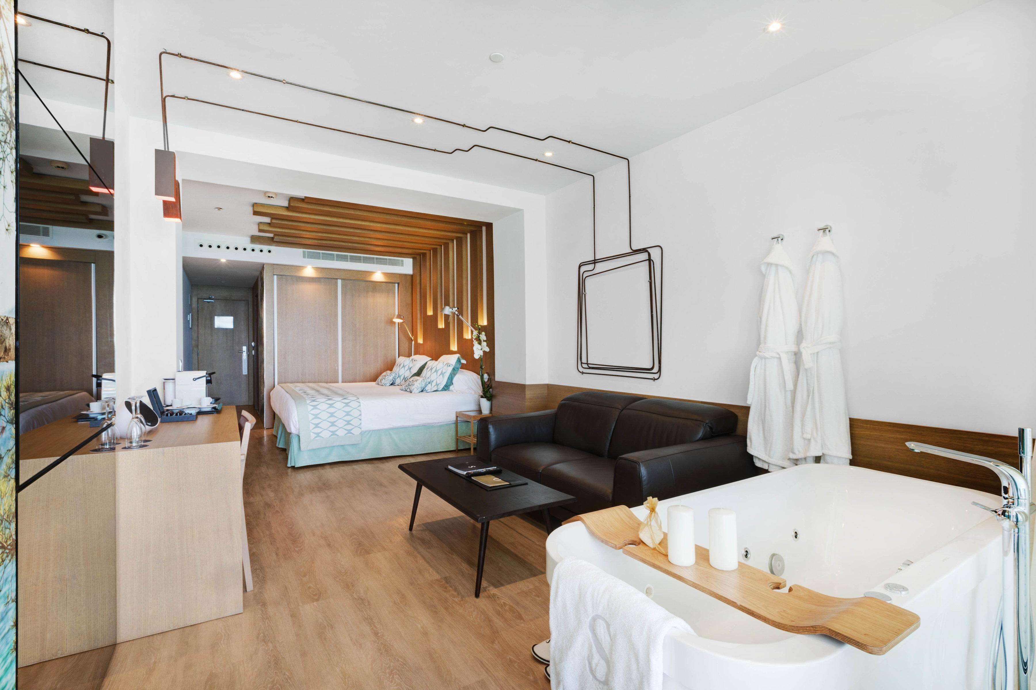Una habitación de hotel moderna con piso de madera, un sofá de cuero negro, una cama con cabecera de madera, una bañera blanca grande, una mesita de noche de madera y un escritorio con espejo y lámpara.