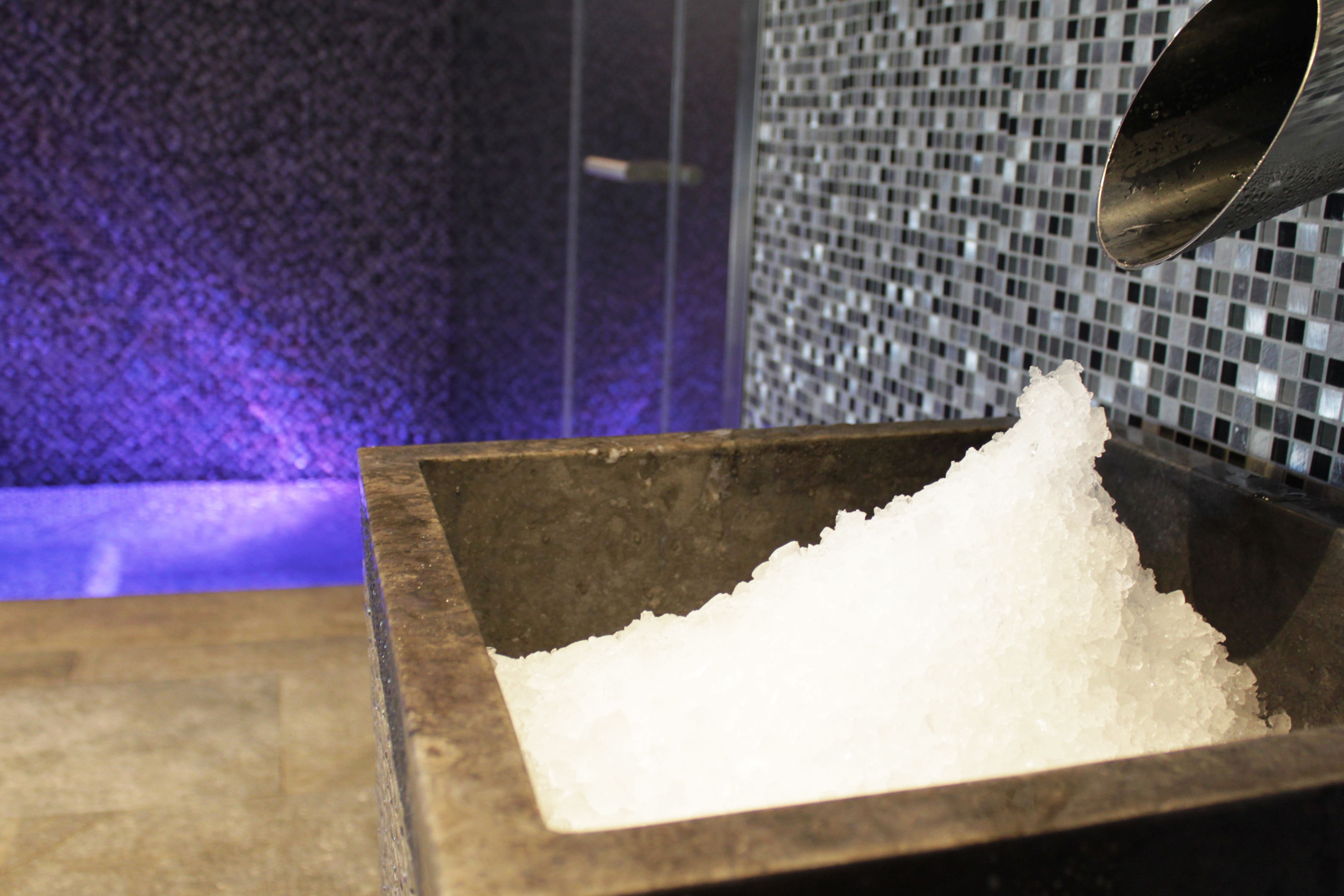 Eine Holzkiste voller Eiswürfel, ein Metallbehälter der Eis ausgießt, und eine geflieste Wand im Hintergrund.
