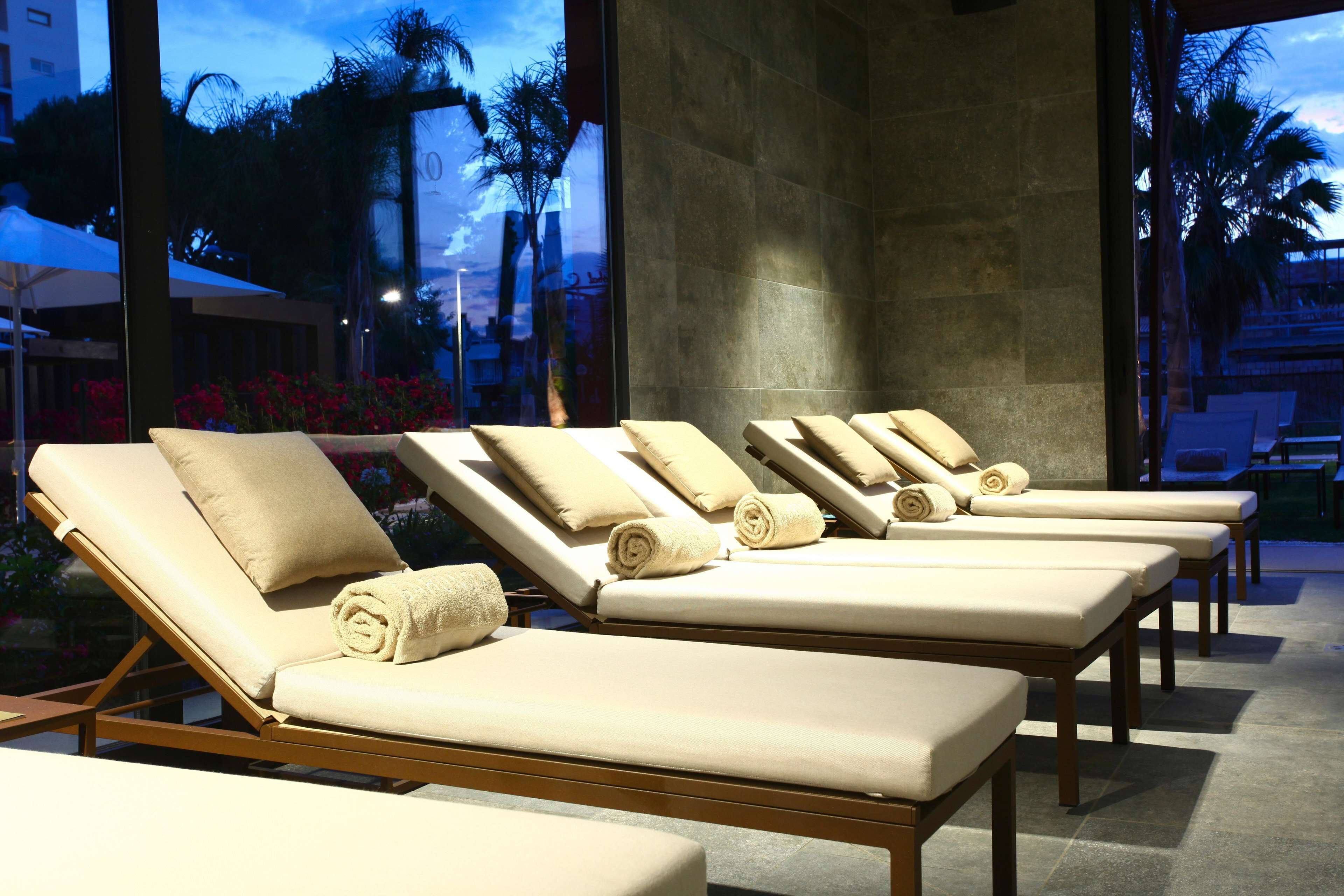 A row of sun loungers with beige cushions and towels, positioned in a modern indoor setting with large windows.