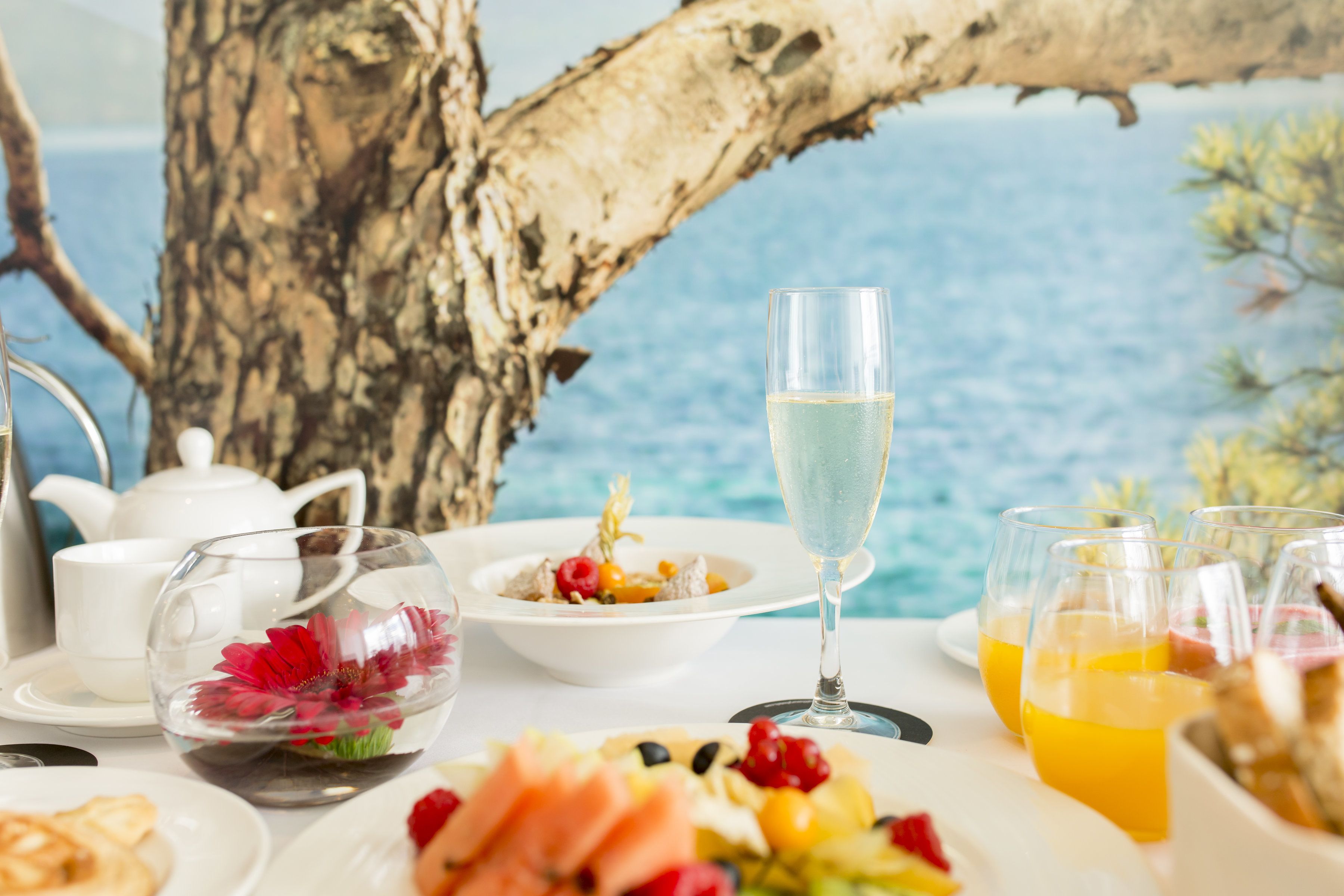 Una mesa dispuesta con una copa de champán, una ensalada de frutas y un bol de comida, con vista al mar de fondo.