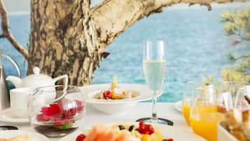 Una mesa dispuesta con una copa de champán, una ensalada de frutas y un bol de comida, con vista al mar de fondo.