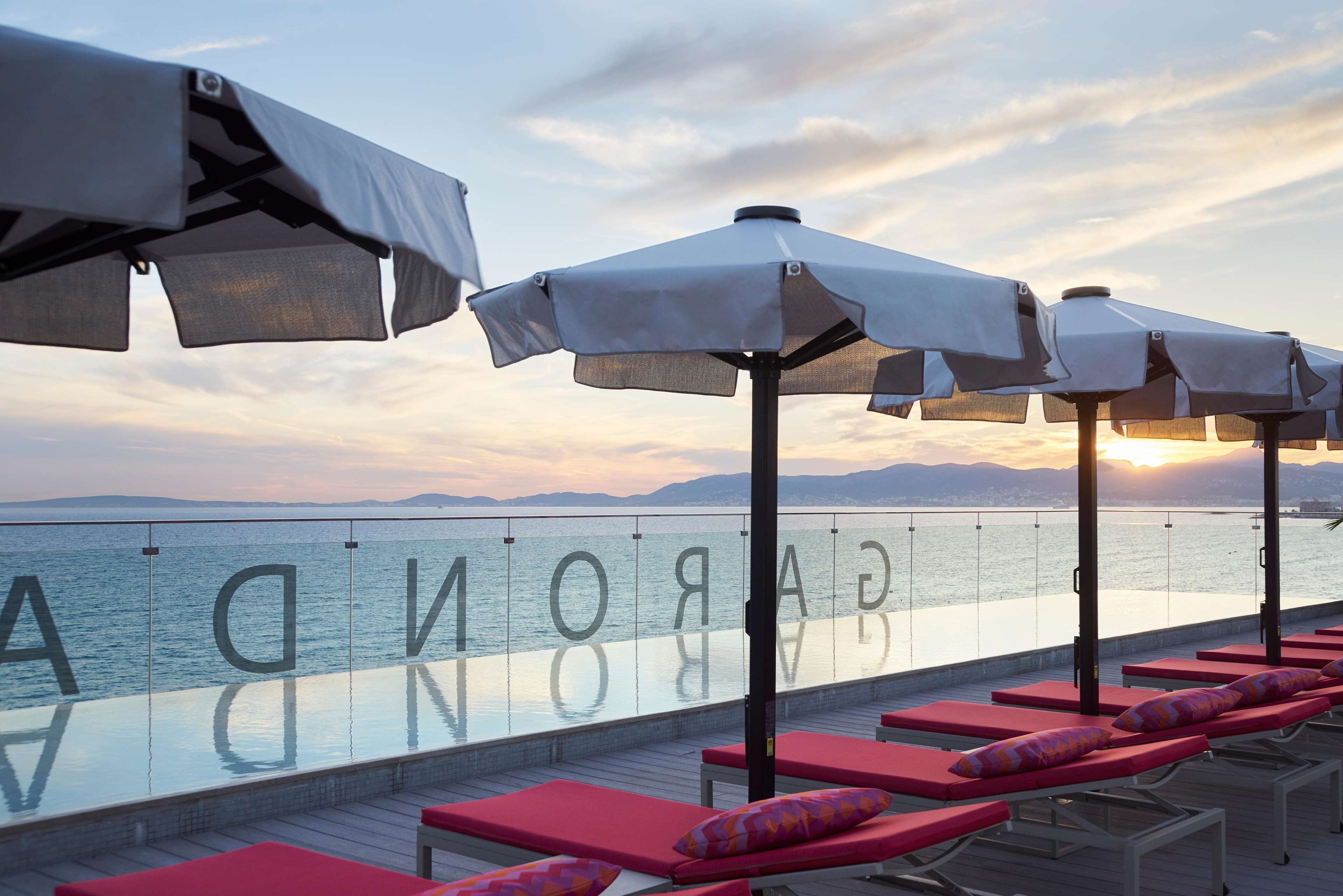 Zona junto a la piscina con sillones de descanso y sombrillas, con vista a un cuerpo de agua y montañas al fondo.