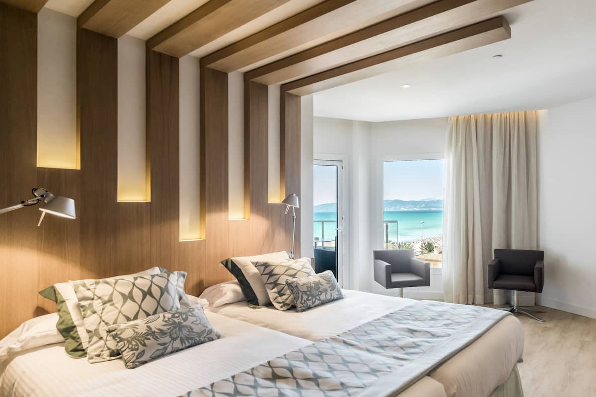 A hotel room with a large bed, two bedside tables, a chair, a desk, and a view of the sea.