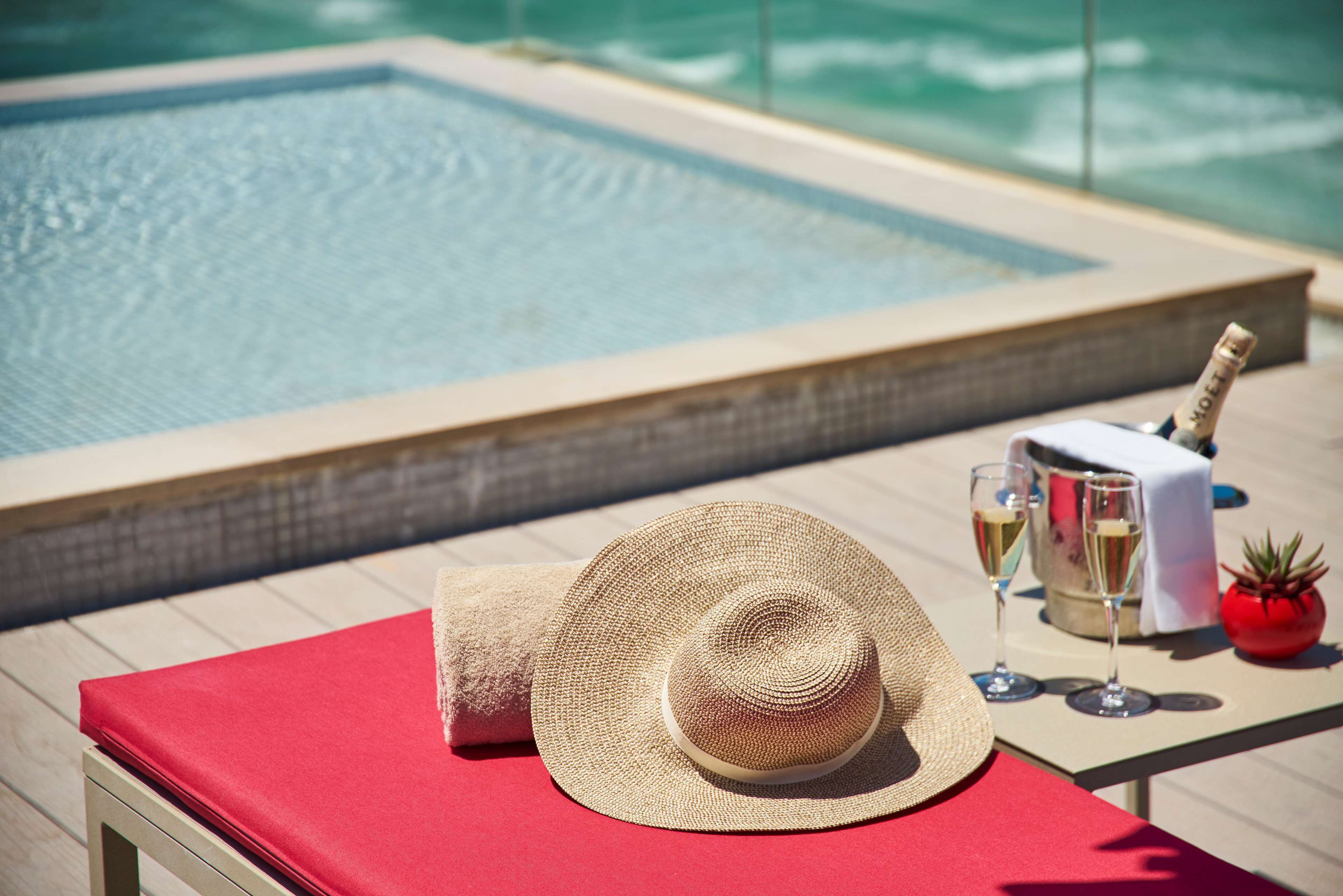 A poolside setting with a red lounge chair, a straw hat, champagne glasses, and a bottle of champagne.