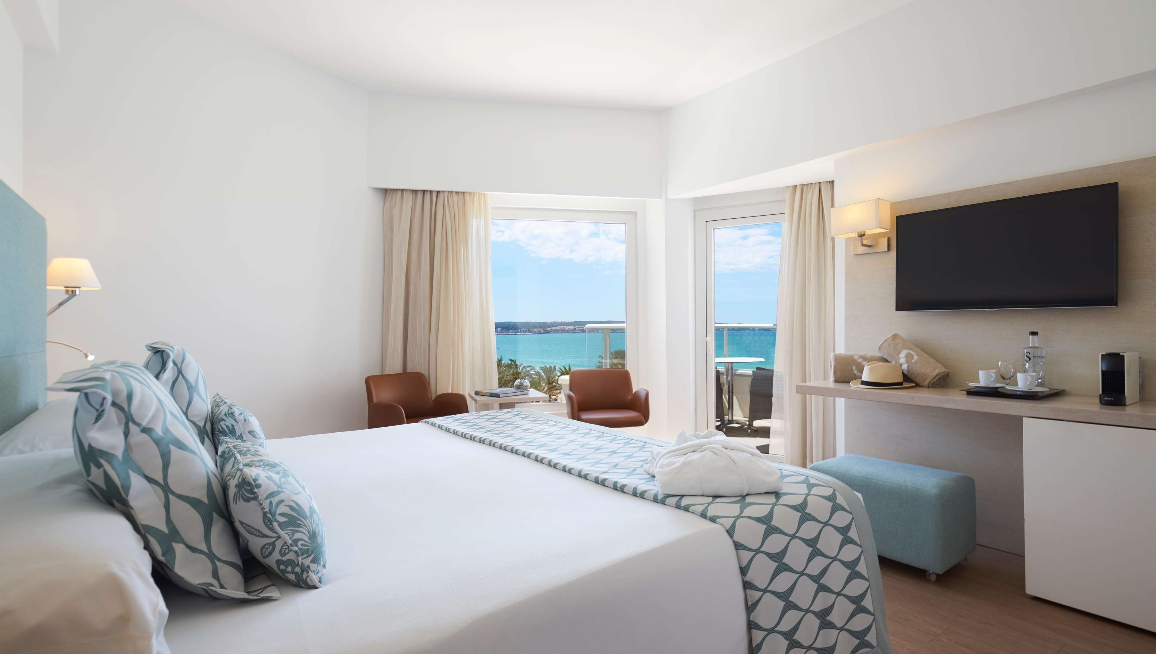 A hotel room with a large bed, a desk, a TV, and a balcony with a view of the sea.