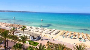 A beach with numerous umbrellas and sun loungers, a clear blue sea, and a few buildings in the background. - Pure Salt Garonda - Adults Only (Palma de Mallorca)