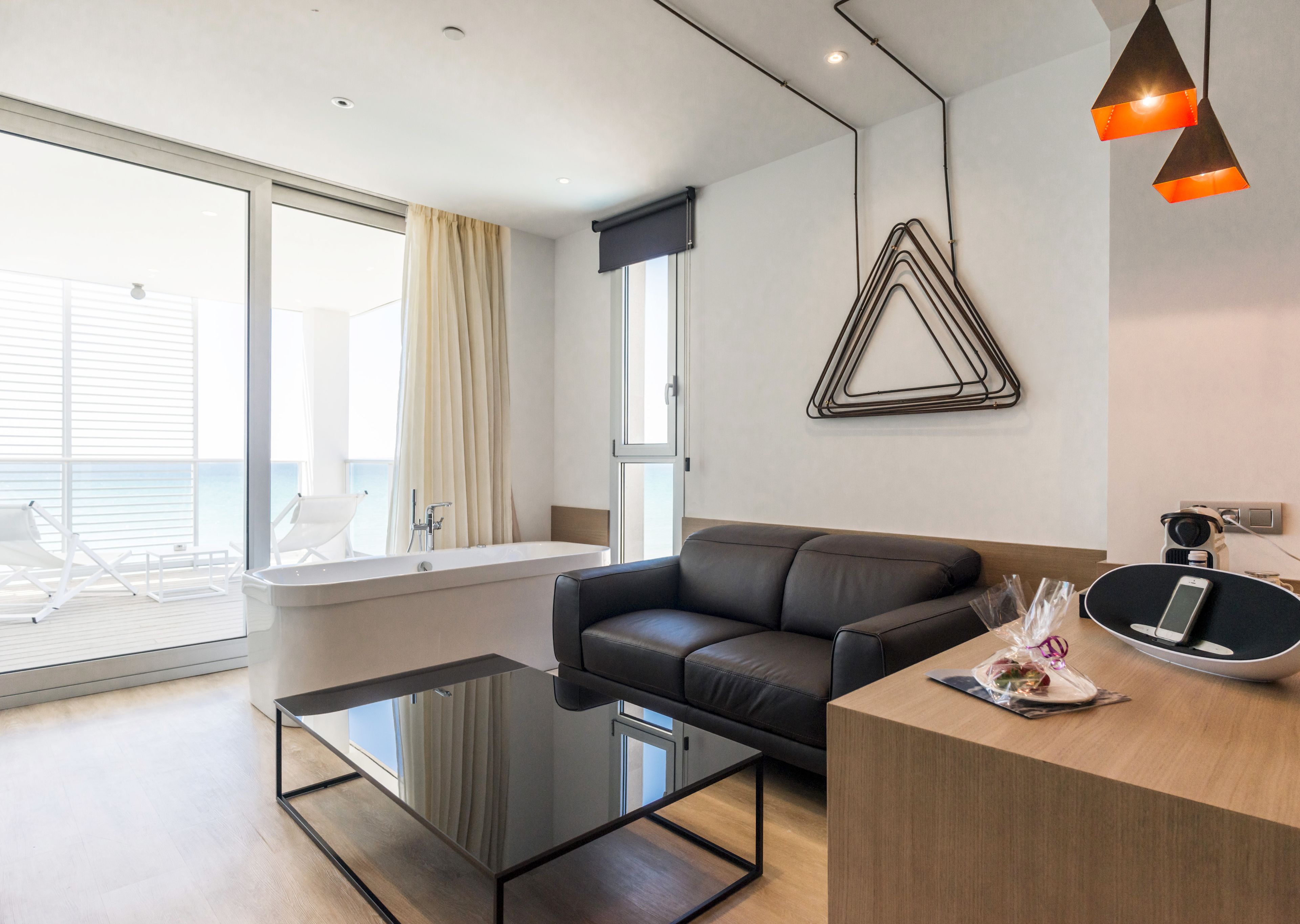 A modern living room with a black leather sofa, a glass coffee table, a wooden side table, and a view of the sea through large windows.