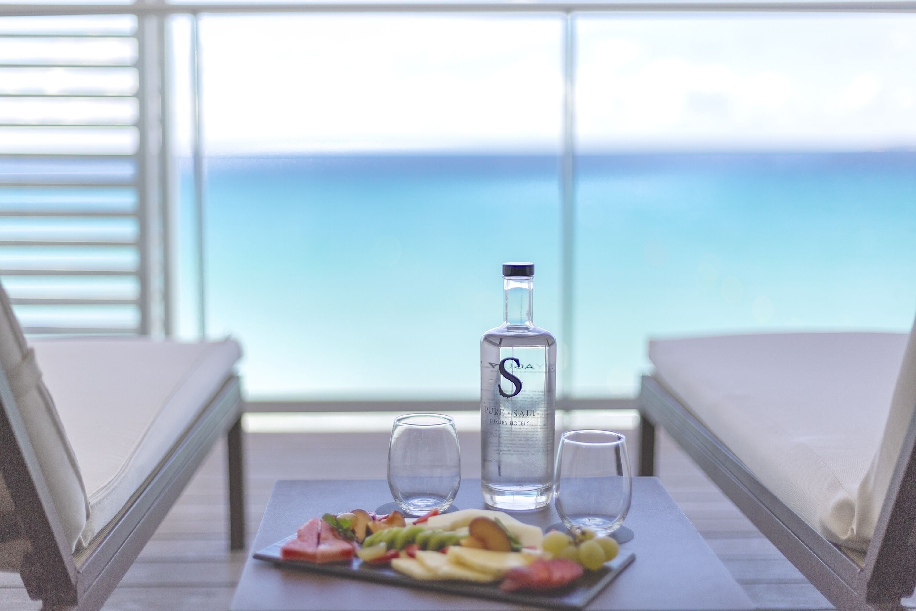 A table with a bottle of water, two glasses, and a fruit platter on a balcony overlooking the ocean.