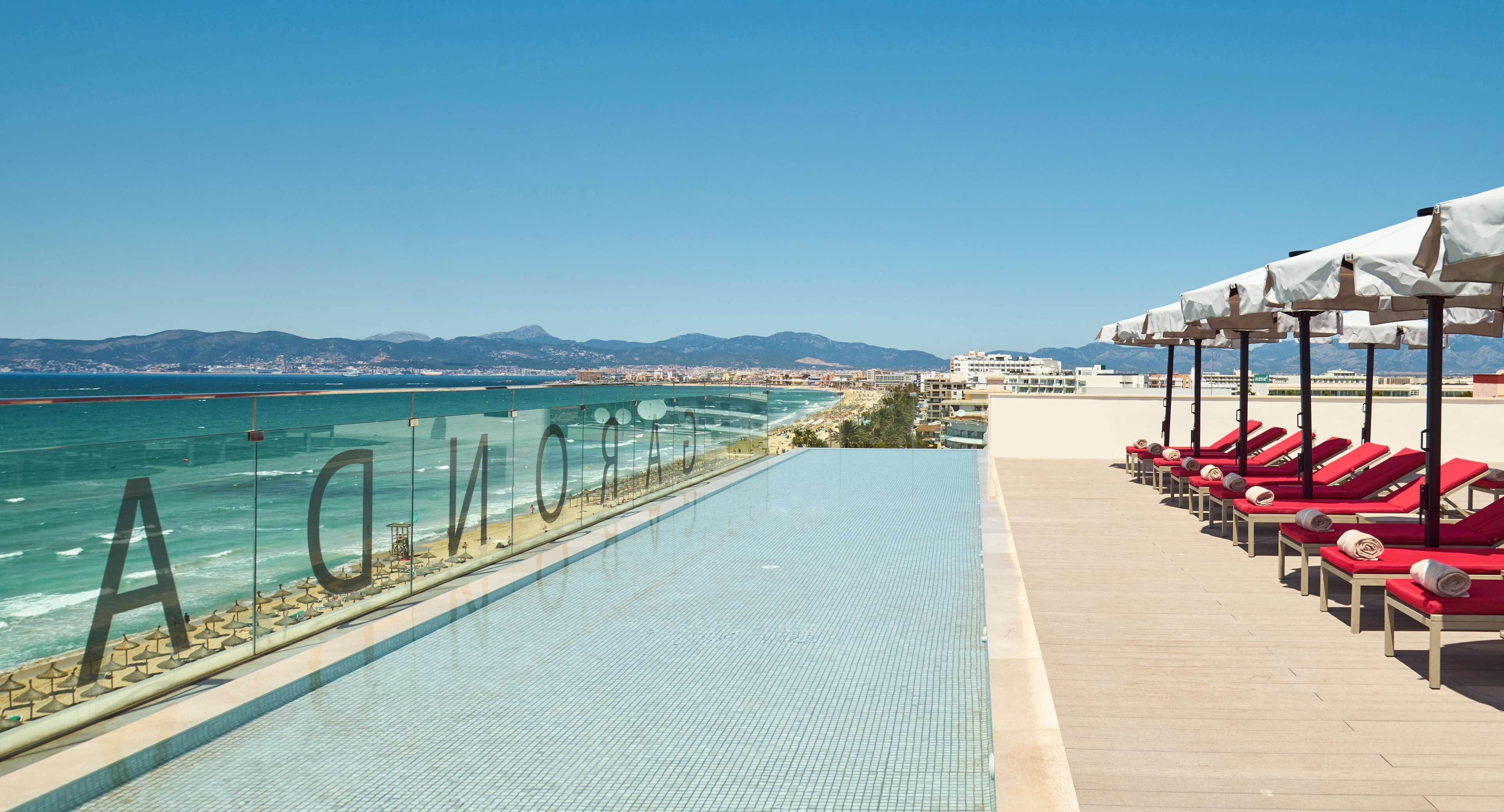 An infinity pool with a view of the sea and mountains, red lounge chairs, and umbrellas.