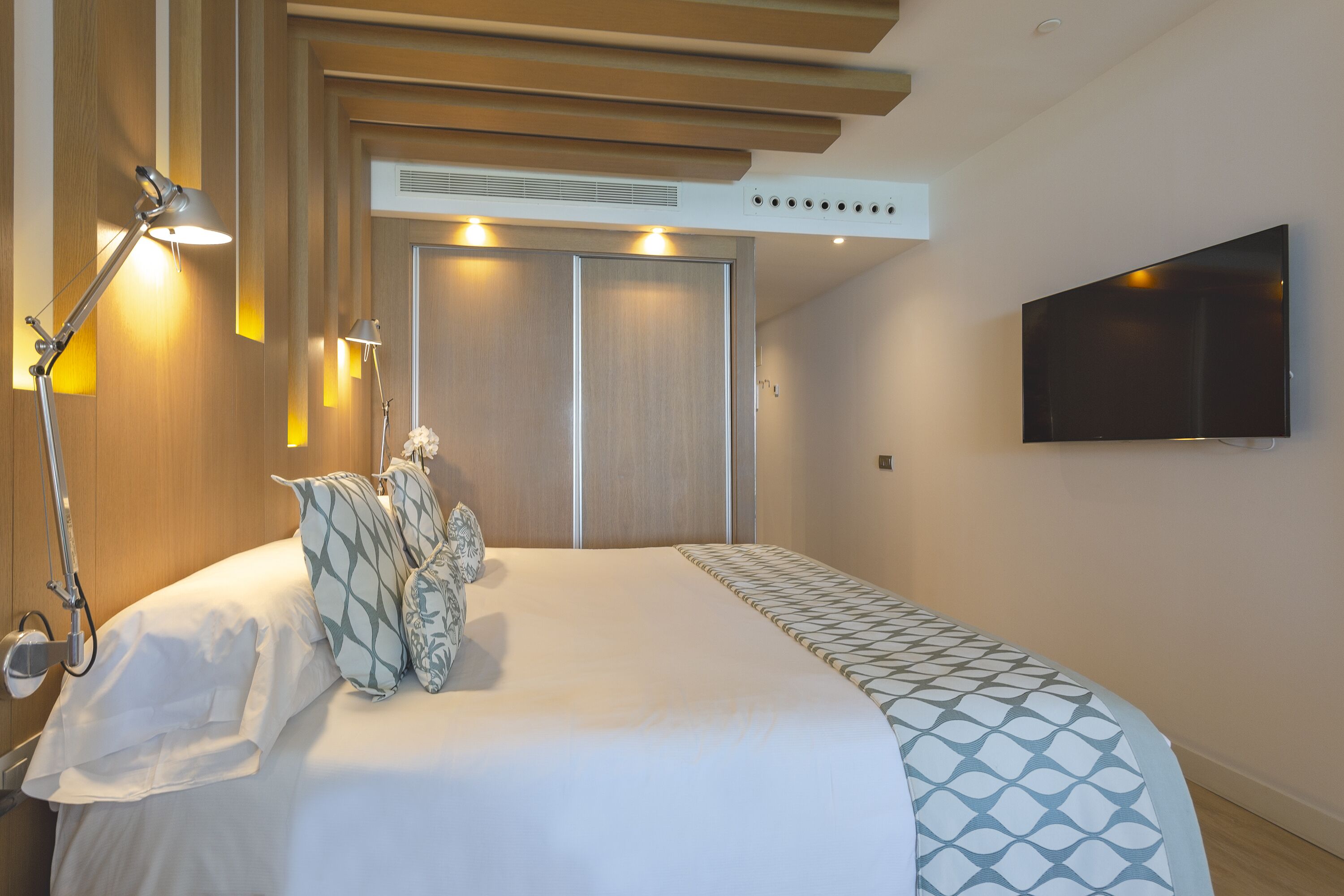 A hotel room with a large bed, a bedside lamp, a TV mounted on the wall, and a wooden wardrobe.