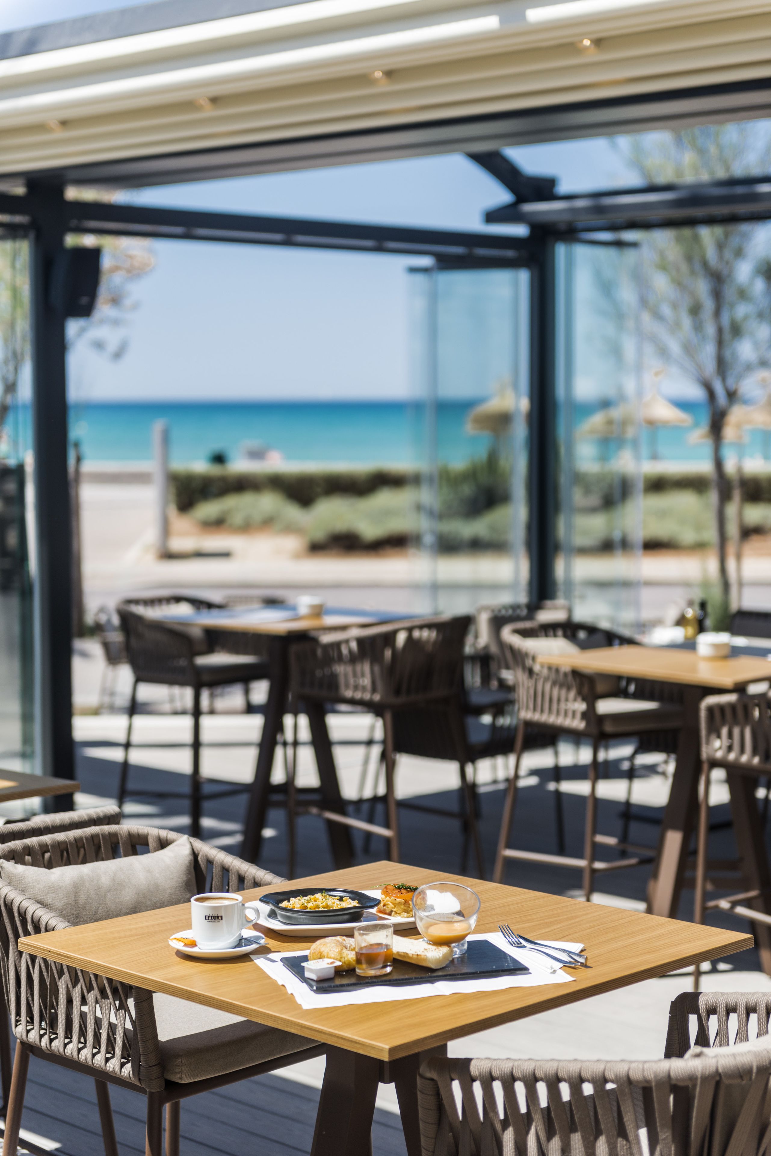 Un área de comedor al aire libre con mesas y sillas, vista a la playa y cielo despejado.
