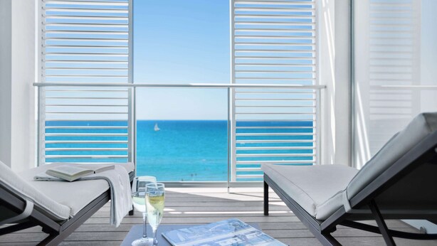 A balcony with two lounge chairs, a table with a magazine, a glass of champagne, and a view of the ocean. - Pure Salt Garonda - Adults Only (Palma de Mallorca)