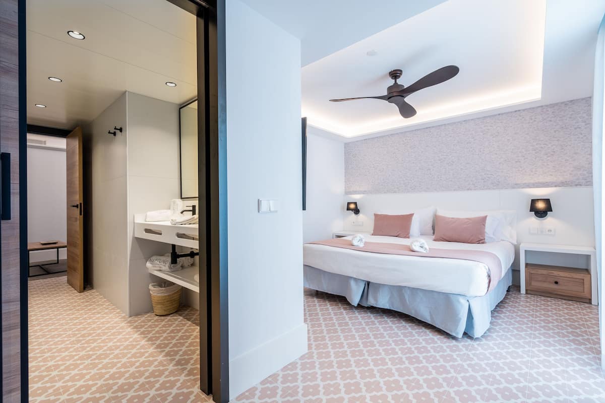 A modern bedroom with a bed, bedside tables, a ceiling fan, and a bathroom with a sink and mirror.