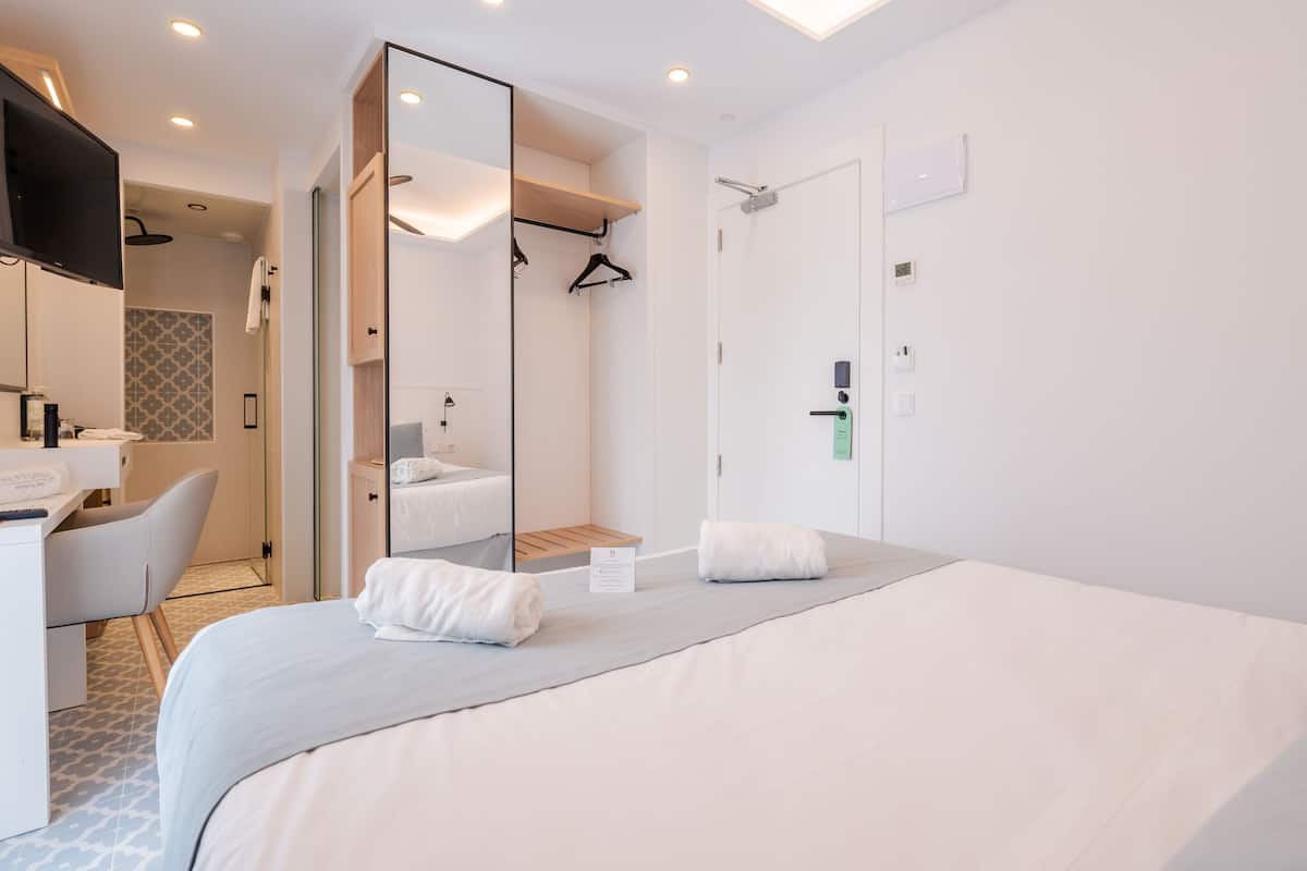 A modern hotel room with a large bed, a desk with a chair, a mirror, a shower area, and a wardrobe.