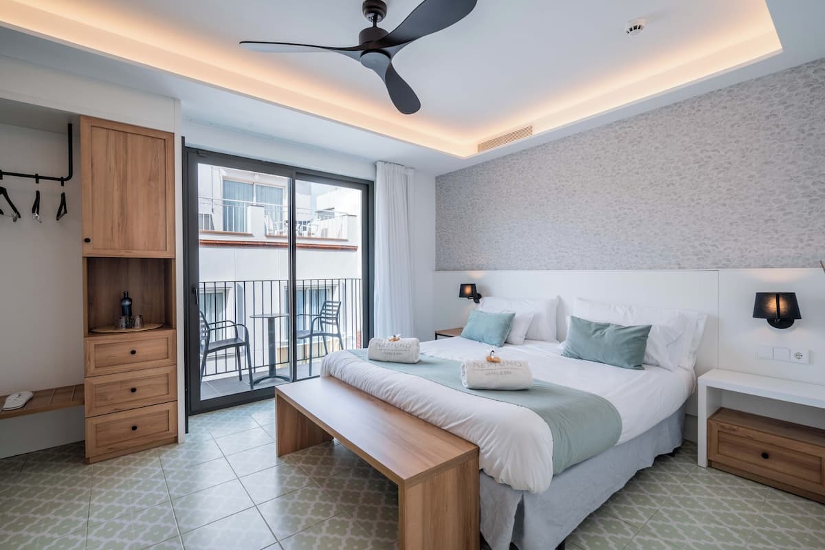 A modern hotel room with a large bed, a wooden bench, a ceiling fan, and a balcony with a view of the city.