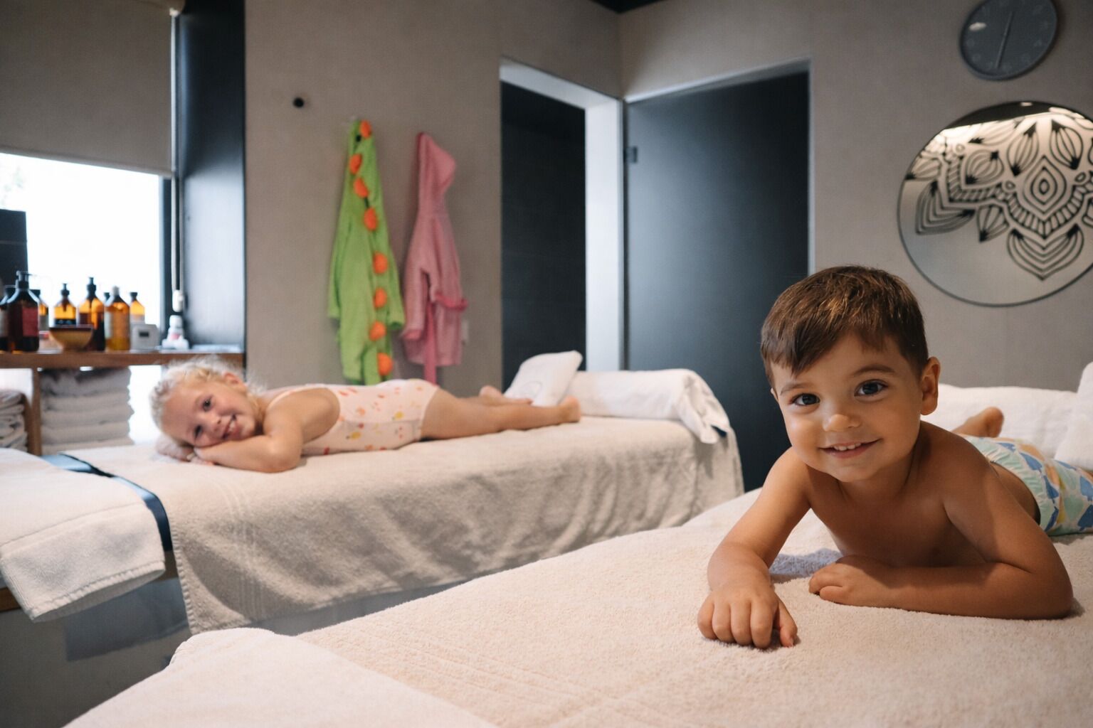 Dos niños recostados en camas dentro de una habitación con ventana, puerta y un reloj de pared.