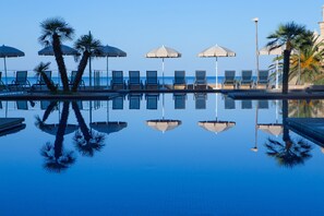 Une piscine à l’eau d’un bleu limpide, entourée de chaises longues et de parasols, dans laquelle se reflètent les palmiers et les chaises.
