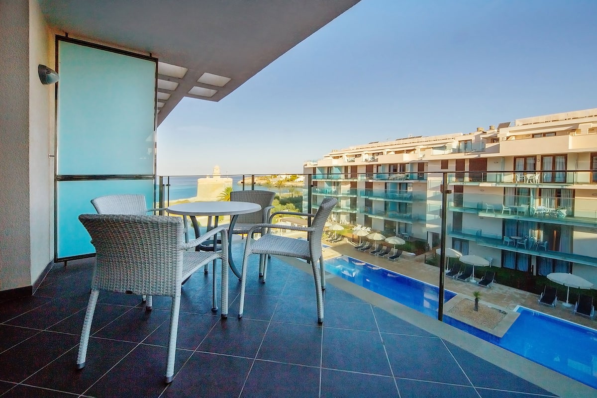 A balcony with a glass railing overlooking a swimming pool and a building with multiple balconies.