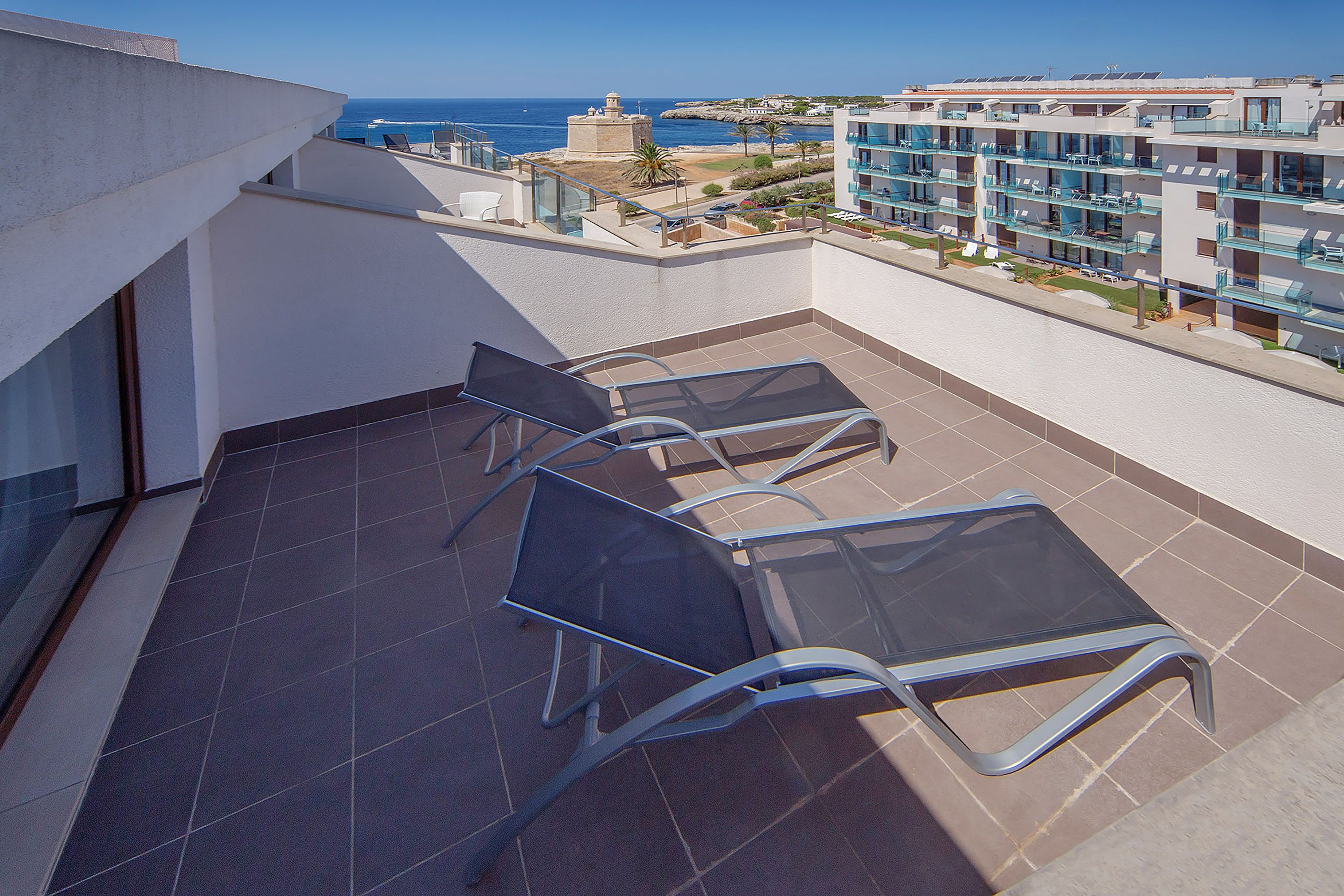 A rooftop terrace with a view of the sea, featuring a seating area with metal chairs and a glass table.