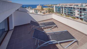 A rooftop terrace with a view of the sea, featuring a seating area with metal chairs and a glass table.