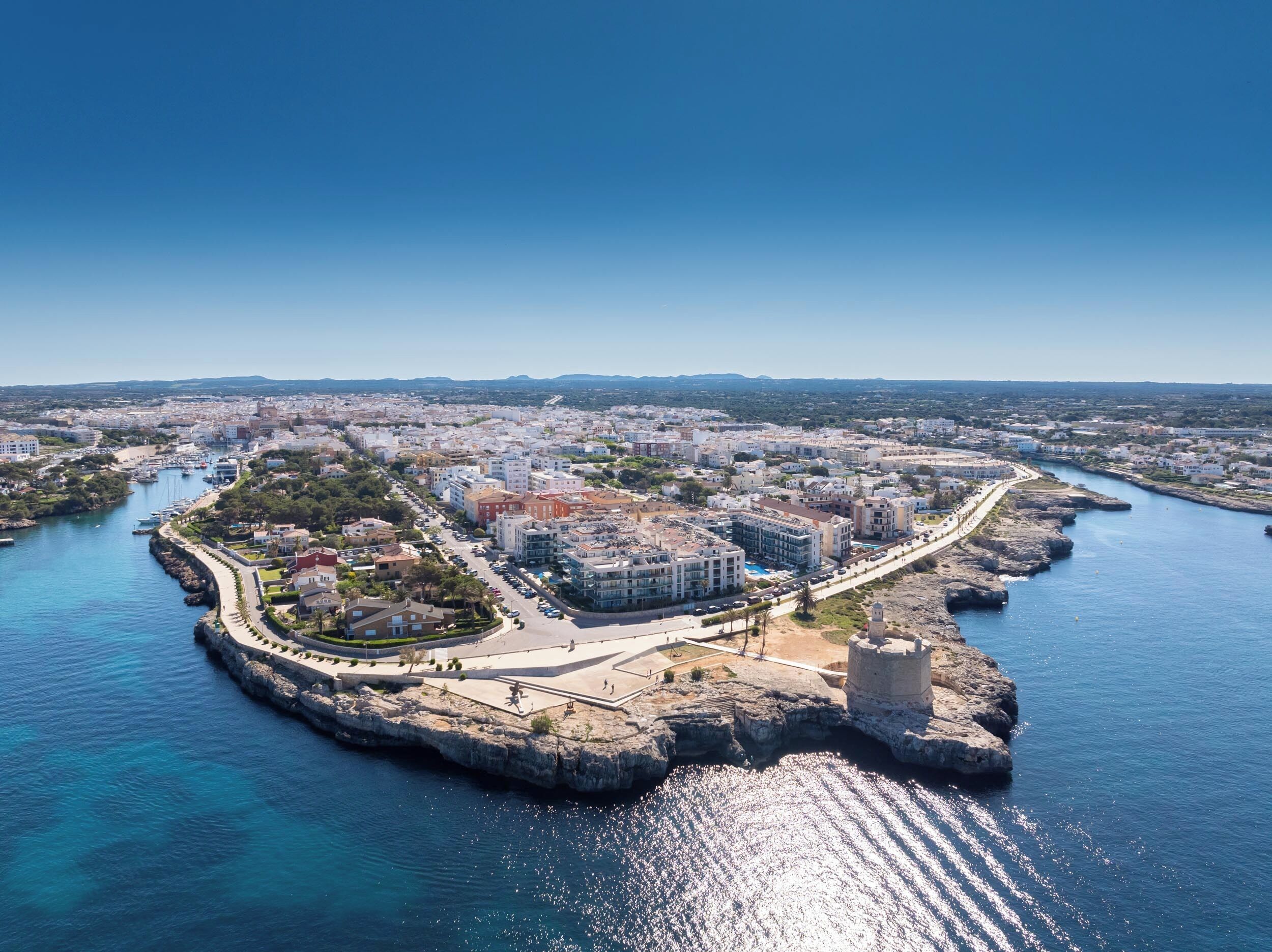 Vista aérea de una ciudad costera con una pronunciada península rocosa, un puerto deportivo y una combinación de edificios residenciales y comerciales.
