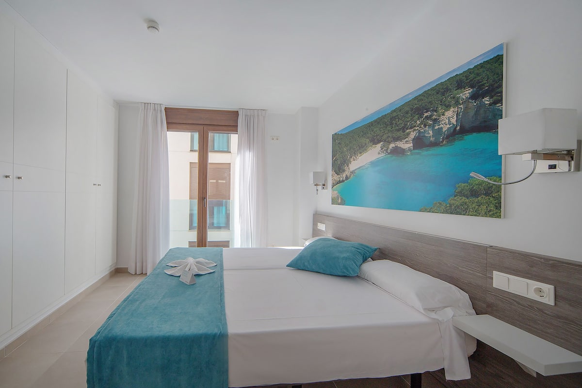 A hotel room with a bed, a nightstand, a painting of a coastal landscape, a window with curtains, and a door.