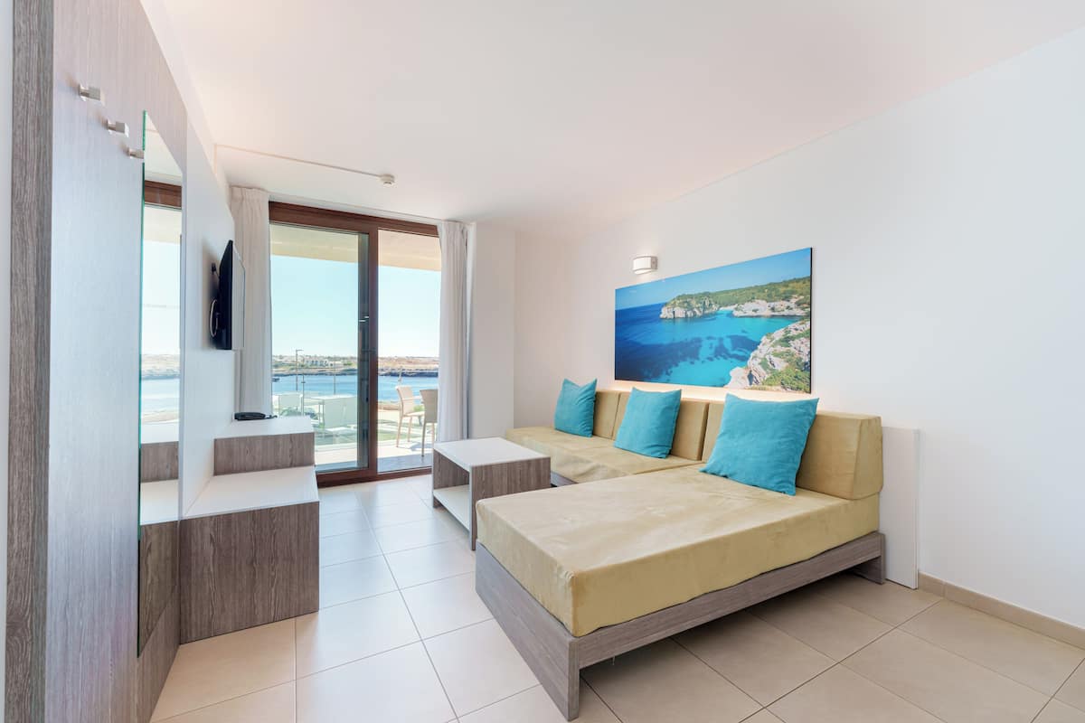 A modern hotel room with a bed, a TV, a balcony with a view of the sea, and a painting of a coastal scene.