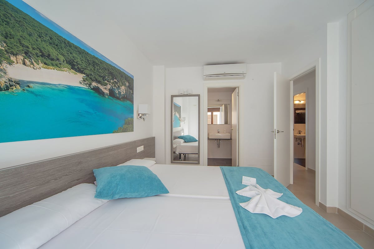 A hotel room with a bed, a large beach mural, a bathroom visible through an open door, and a view of the beach.