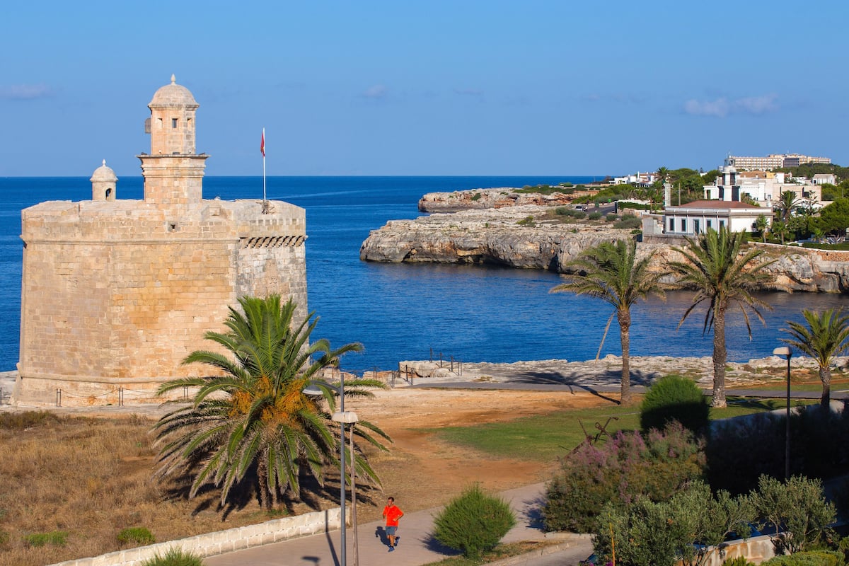 A coastal view with a historic stone tower, a palm tree, and a paved path leading to a building with a red roof.