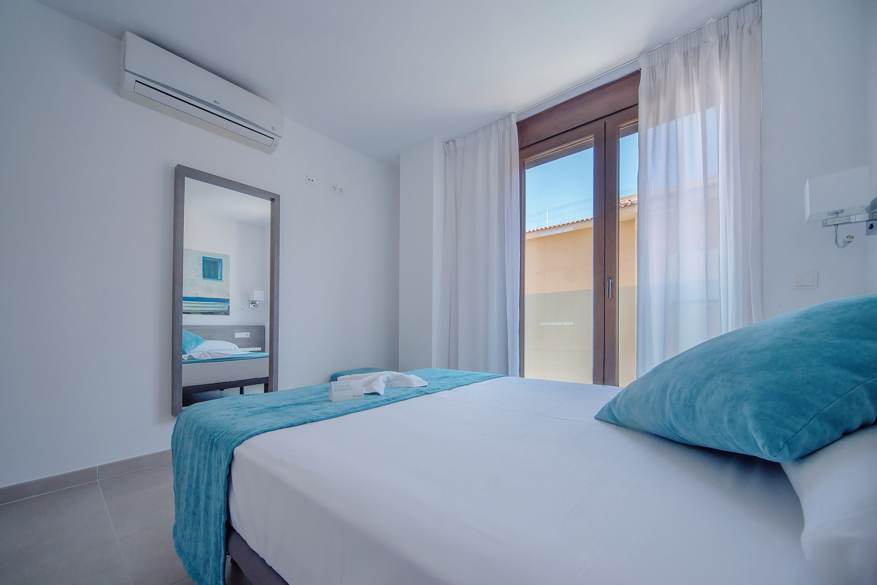 Una habitación de hotel con cama, espejo, ventana con cortinas y un equipo de aire acondicionado.