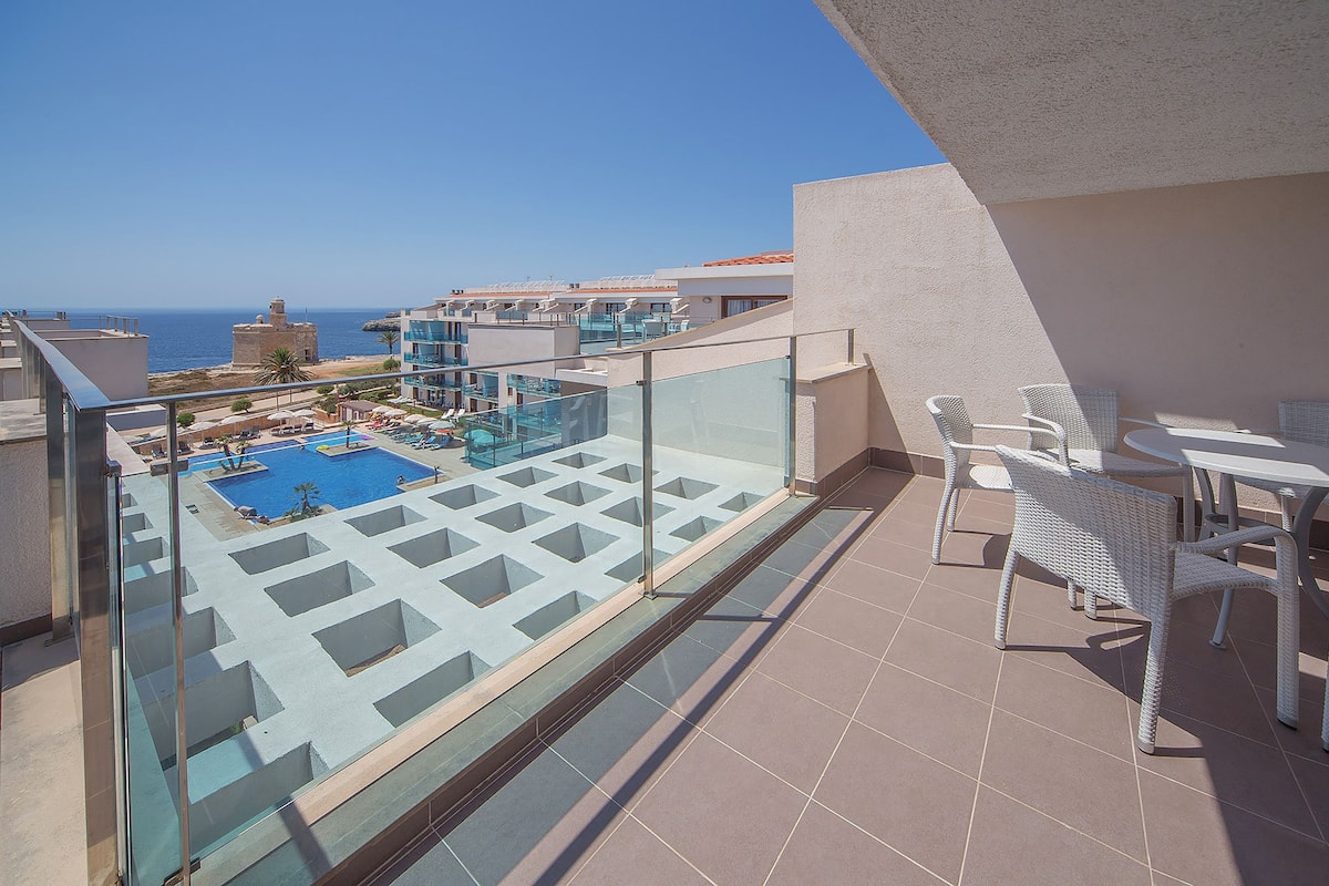 A balcony with a glass railing overlooking a swimming pool and the sea.