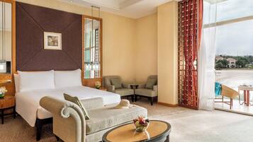 A hotel room with a large bed, a sofa, a small table with fruit, a mirror, and a view of the outside.