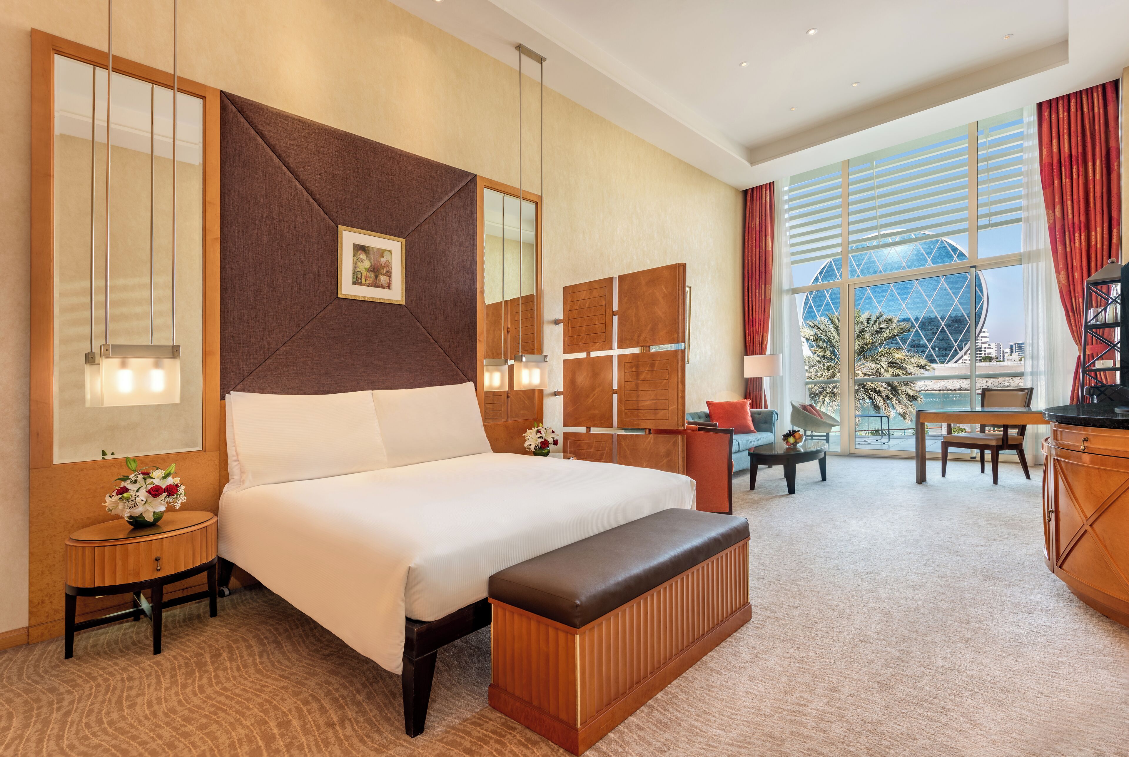 Habitación de hotel con una cama grande, cabecera de madera, mesita de noche con un florero, una banca, un escritorio y un ventanal con vistas a palmeras y edificios.