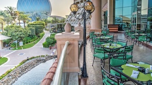 Een hotel met terras, een groot glazen gebouw op de achtergrond en een verzorgde tuin.
