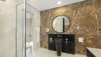 Une salle de bain moderne dotée d’une douche à parois de verre, d’un mur en marbre, d’un miroir rond et d’un lavabo noir avec un receveur en verre.