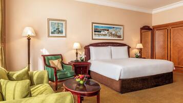 A hotel room with a large bed, two chairs, a table with fruit, and two paintings on the wall.